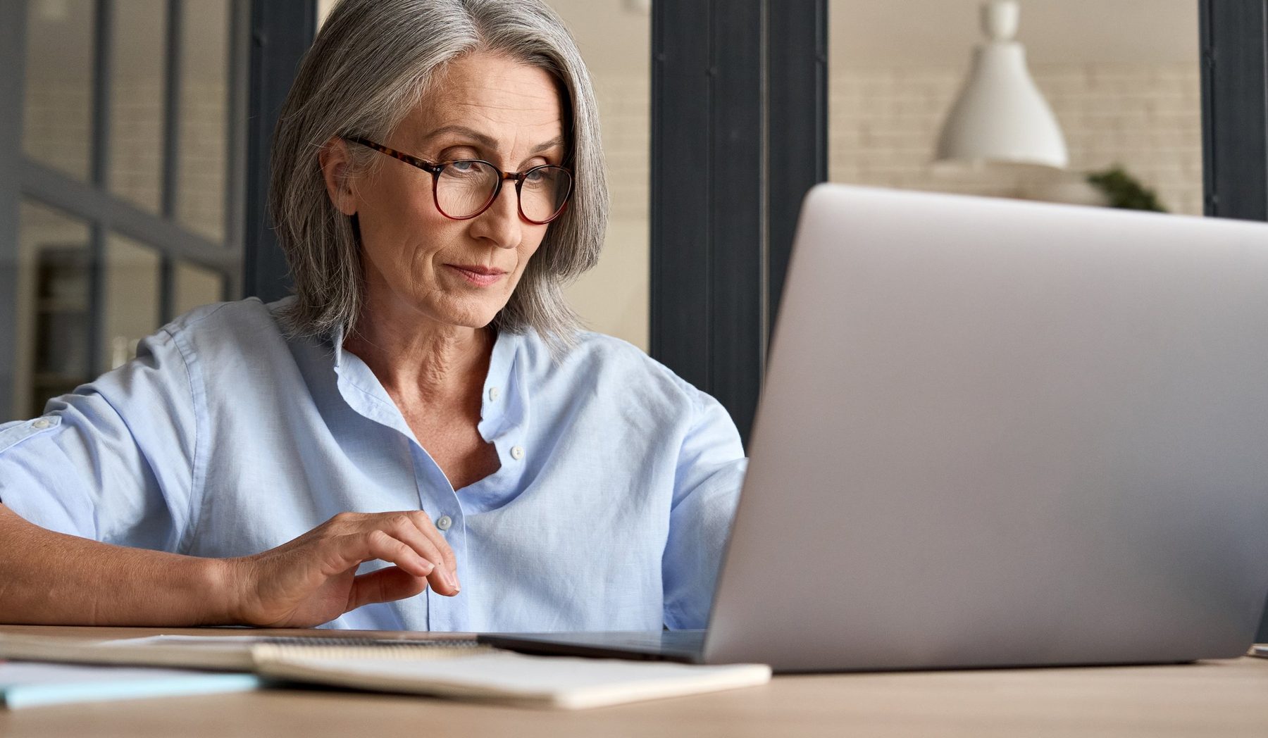 An older woman with glasses working on a laptop at a desk